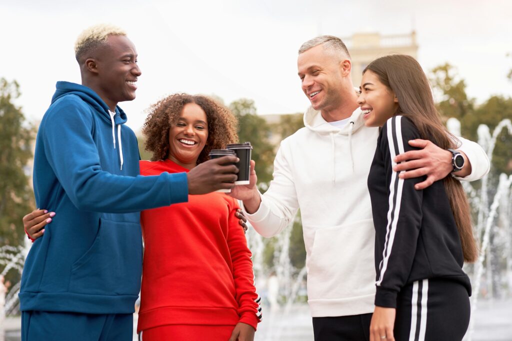 Cheerful diverse friends toasting coffee cups in park after workout