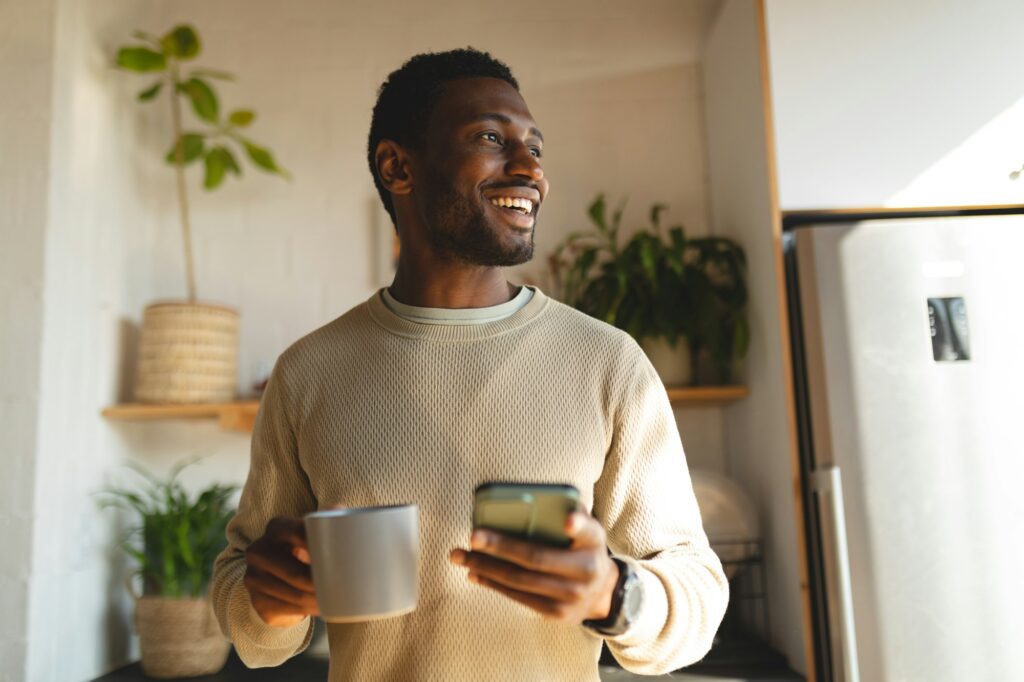 Happy african american man using smartphone and drinking coffee in kitchen