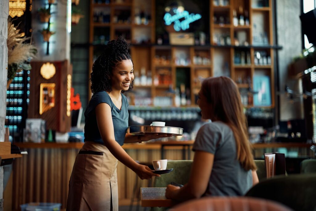 Happy black waitress serving coffee to a guest in cafe.