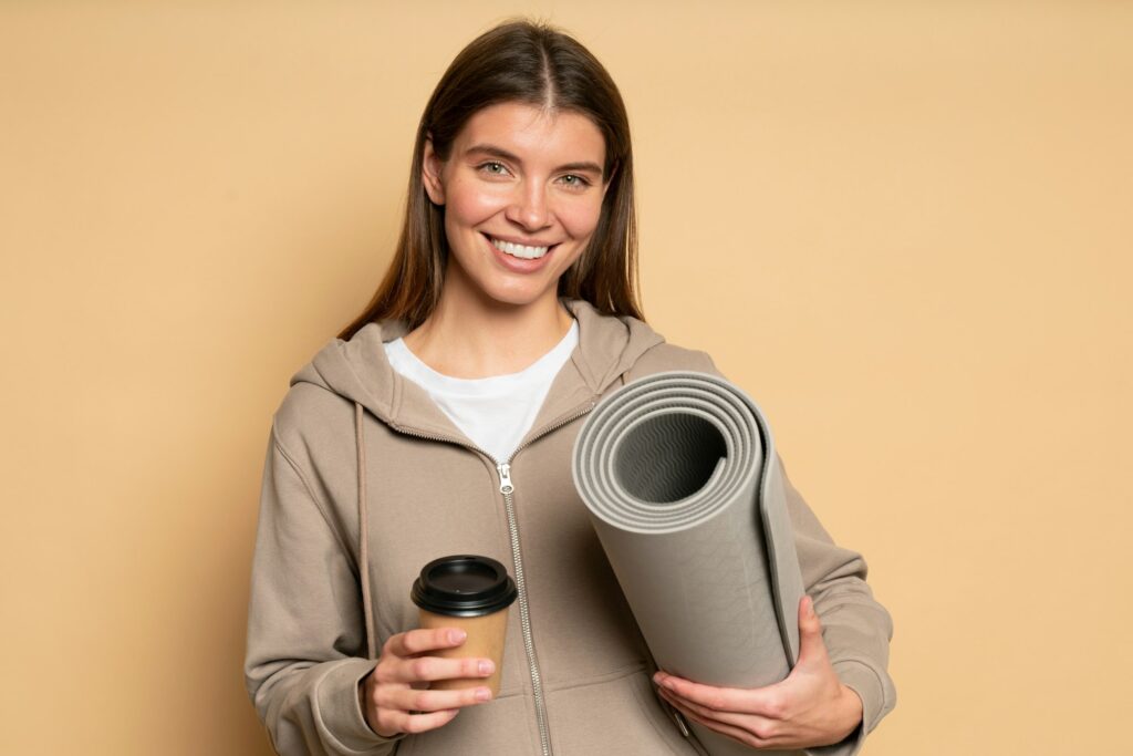 Woman fitness instructor posing after workout with rolled mat and plastic cup of coffee